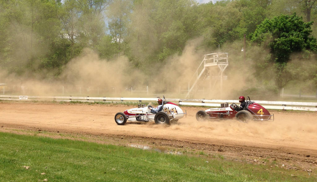 Open-wheel midget race cars sliding through a dirt oval, kicking up clay